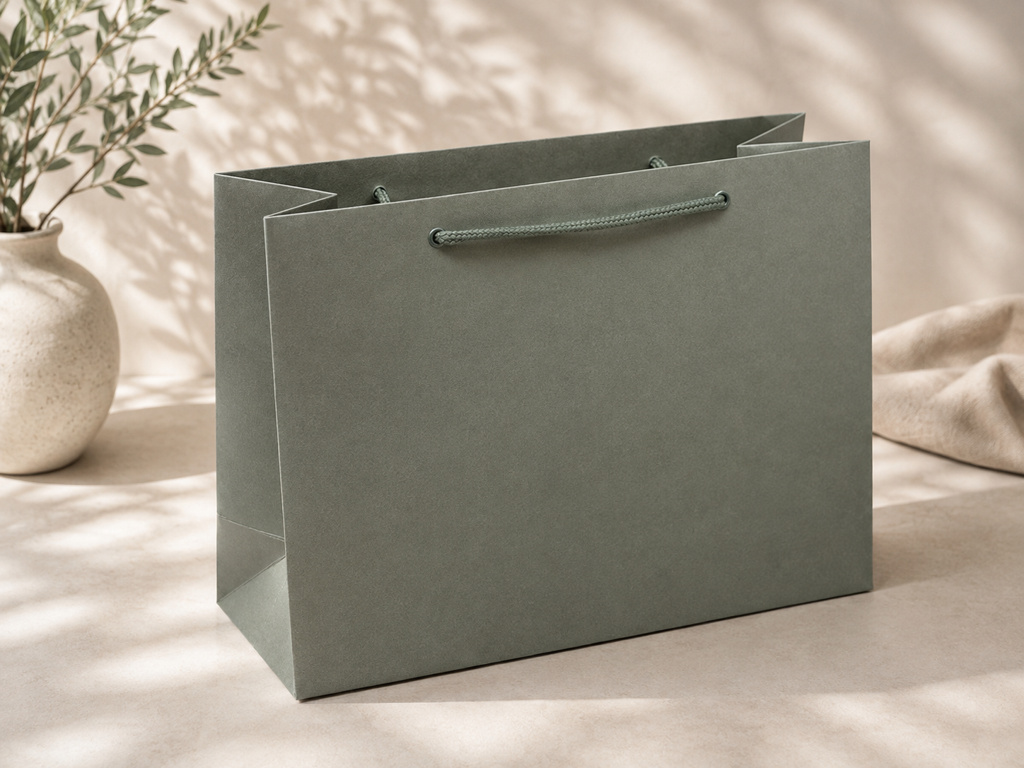 Eine große, mattgrüne „Geschenk-Tragetasche Pure, Größe L, verschiedene Farben“ von Artoz Papier AG mit stabilen Henkeln liegt auf einer beigen Fläche. Im sanft beleuchteten Hintergrund erscheinen eine Keramikvase und ein Stück Stoff verschwommen, während sanftes Tageslicht dezente Schatten wirft – was diese elegante Geschenktüte zur perfekten Wahl für jeden besonderen Anlass macht.