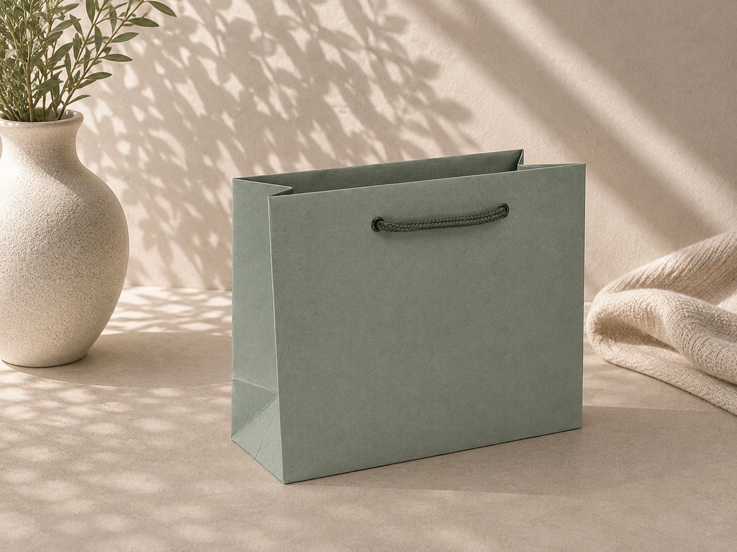 Un « sac cadeau Pure M » vert de la marque Artoz Papier AG, doté de poignées foncées, repose sur une surface beige à côté d'un vase texturé rempli de feuilles vertes, éclairé par une douce lumière du soleil et des ombres délicates en arrière-plan.