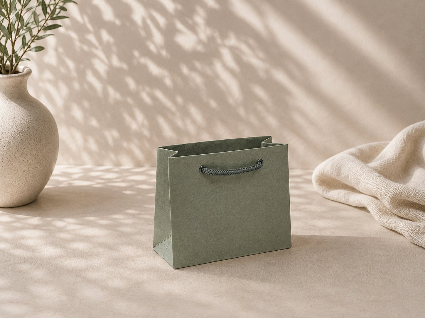 Eine Geschenktüte „Pure“ der Marke Artoz Papier AG in Größe S in Grün mit Kordelgriffen liegt auf einer beigen Fläche neben einer Keramikvase mit Blättern und einem strukturierten Tuch, beleuchtet von sanftem, natürlichem Sonnenlicht.