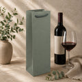 Un sac à vin « Artoz Papier AG » de différentes couleurs, ici en vert, est posé à côté d'une bouteille de vin rouge, d'un verre à vin rempli, d'un vase contenant des plantes vertes et d'un tissu beige, le tout sur une surface beige texturée.