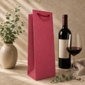Eine Wein-Tragetasche in verschiedenen Farben von Artoz Papier AG, gefertigt aus Kraftpapier, steht neben einer Flasche Rotwein, einem gefüllten Weinglas, einer Vase mit grünem Laub und einem beigen Tuch auf einer in neutralen Farbtönen gehaltenen Fläche, auf die Sonnenflecken fallen – eine stilvolle Geschenkverpackung und praktische Tasche für jeden Anlass.