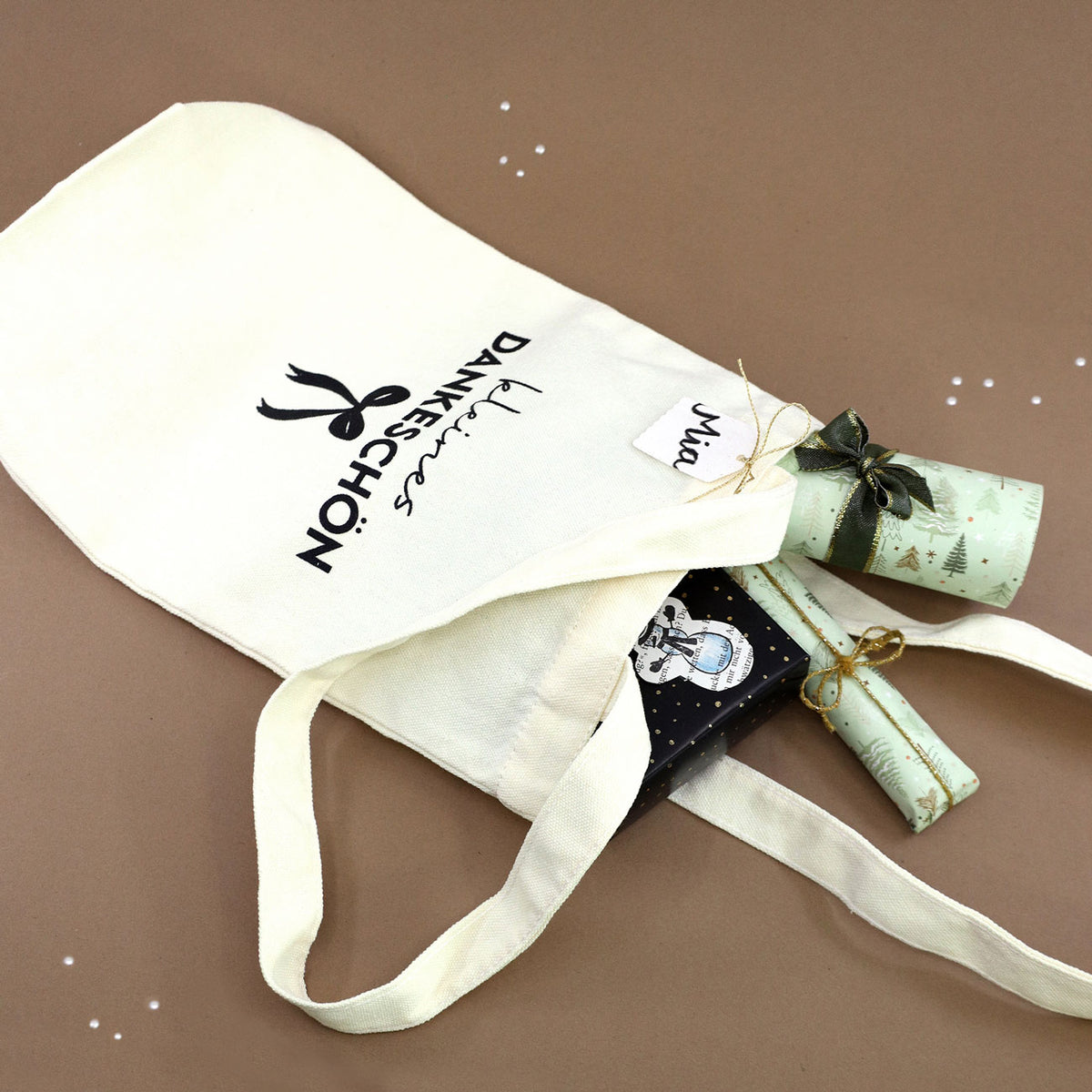 Un sac cadeau en tissu de couleur crème, orné du motif « Petit merci » de la marque Artoz Papier AG, contient trois cadeaux emballés et une étiquette portant le nom « Nia », le tout posé sur un fond marron parsemé de petits points décoratifs blancs : un emballage écologique idéal pour tout cadeau de remerciement.