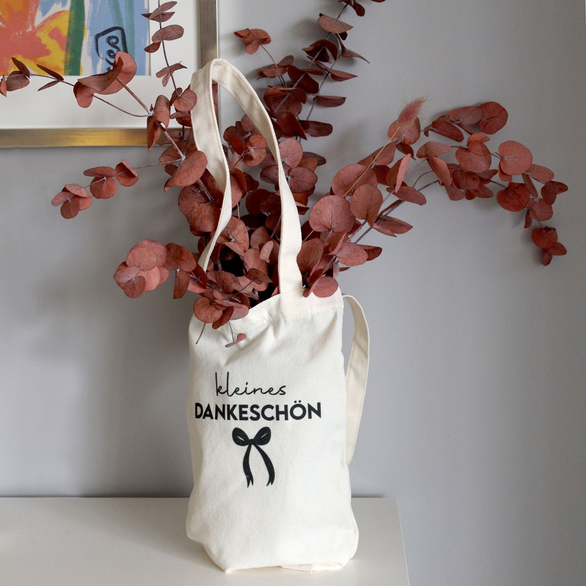 Un sac-cadeau en tissu de couleur crème orné d'un motif « Kleines Dankeschön » d’Artoz Papier AG, ornée d’un nœud imprimé et remplie de brindilles d’eucalyptus séchées de couleur brun-rouge, repose sur une table blanche devant un mur gris : un emballage écologique, idéal comme emballage cadeau réutilisable pour toutes les occasions.