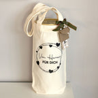 Un sac cadeau en tissu couleur crème de la société Artoz Papier AG, orné du motif « Von Herzen für Dich » (Du fond du cœur, pour toi), est décoré de cœurs noirs et d'un texte en allemand ; fermé par un ruban vert et des étiquettes, il se détache nettement sur le fond clair – un emballage écologique, idéal pour présenter du vin avec élégance.