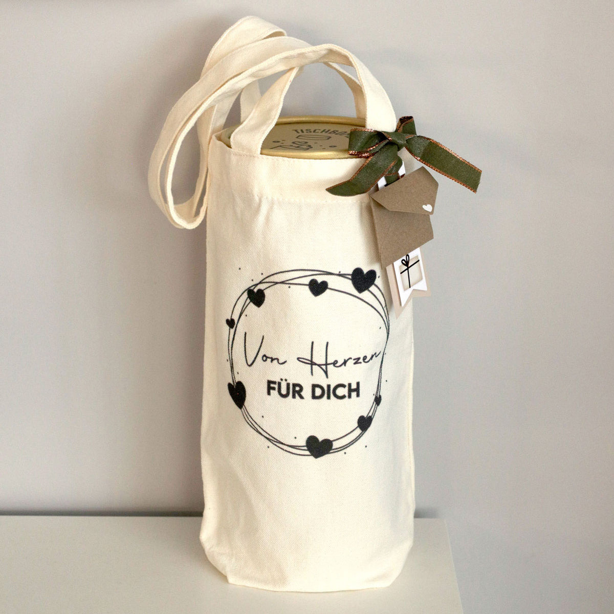 Un sac cadeau en tissu couleur crème de la société Artoz Papier AG, orné du motif « Von Herzen für Dich » (Du fond du cœur, pour toi), est décoré de cœurs noirs et d'un texte en allemand ; fermé par un ruban vert et des étiquettes, il se détache nettement sur le fond clair – un emballage écologique, idéal pour présenter du vin avec élégance.