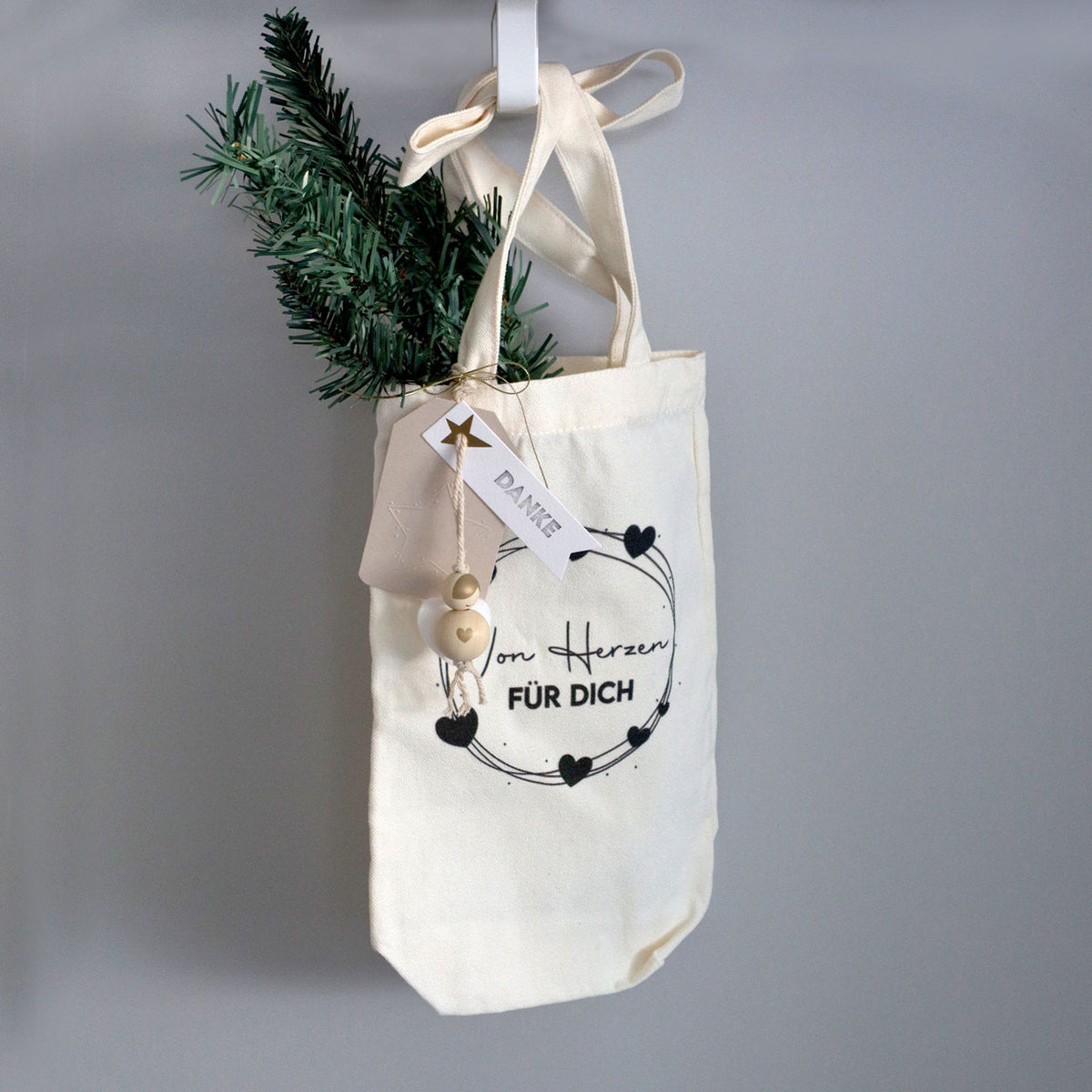 Un sac-cadeau en tissu de couleur crème, orné du motif « Von Herzen für Dich » de la marque Artoz Papier AG et décoré de cœurs noirs, est suspendu à un crochet. Un petit rameau de pin, une décoration blanche et une étiquette portant l'inscription « Danke » sont fixés au sac, ce qui en fait un emballage charmant et écologique pour votre cadeau spécial.