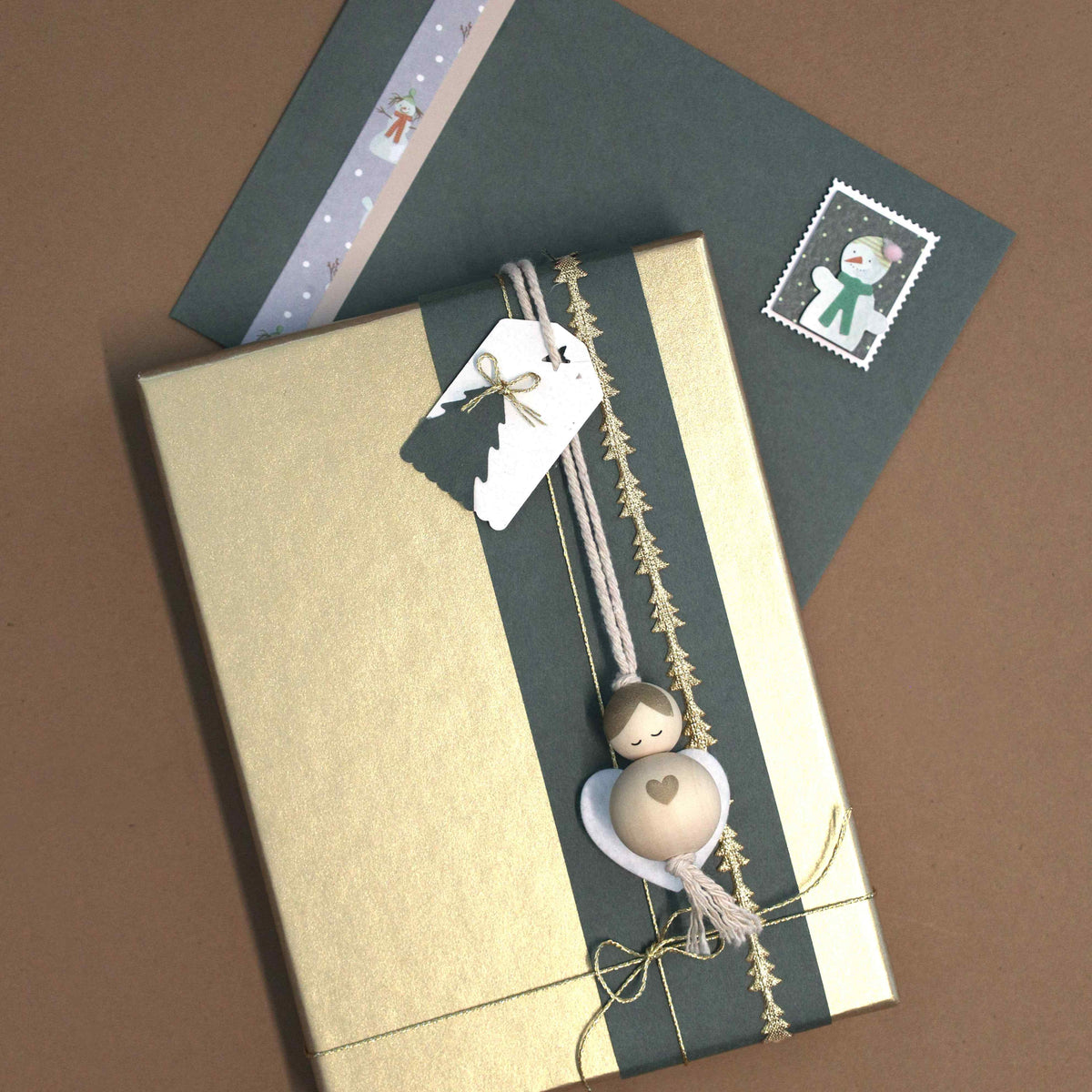 Un cadeau emballé dans du papier vert et doré, orné d'une décoration en bois « Artoz Papier AG Geschenkanhänger Engel », repose à côté d'une enveloppe verte, agrémentée d'un autocollant représentant de la neige et d'un timbre-poste, le tout disposé sur un fond marron.