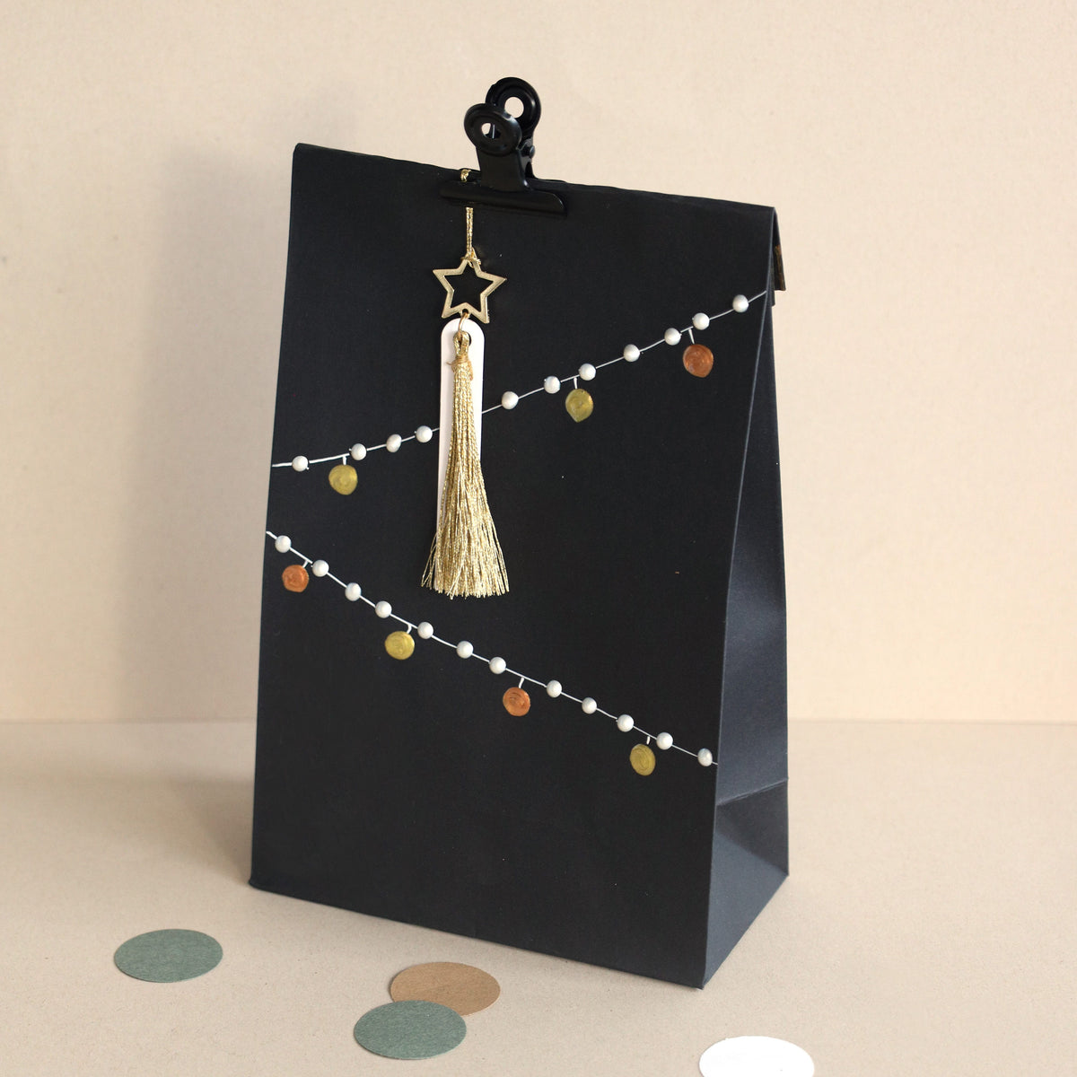 Un sac-cadeau en papier noir est orné d'une ficelle blanche et de petits points colorés ; il est fermé par une pince noire et agrémenté d'un pompon doré ainsi que de l'étiquette « Geschenkanhänger Stern » d'Artoz Papier AG, ornée d'un pendentif en forme d'étoile. Les étiquettes « Kraft Anhänger » apportent une touche charmante, idéale pour les projets de bricolage. À proximité, des découpes circulaires en papier sont dispersées sur la surface beige.