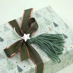 Un cadeau emballé dans du papier bleu clair orné de motifs d'arbres, noué avec un ruban vert et cuivré, agrémenté d'un pompon en fil vert et d'une petite breloque en forme d'étoile argentée, est parfaitement mis en valeur par l'étiquette « Geschenkanhänger Tannenbaum » d'Artoz Papier AG — idéale pour apporter cette touche spéciale « DIY-Projekte ».