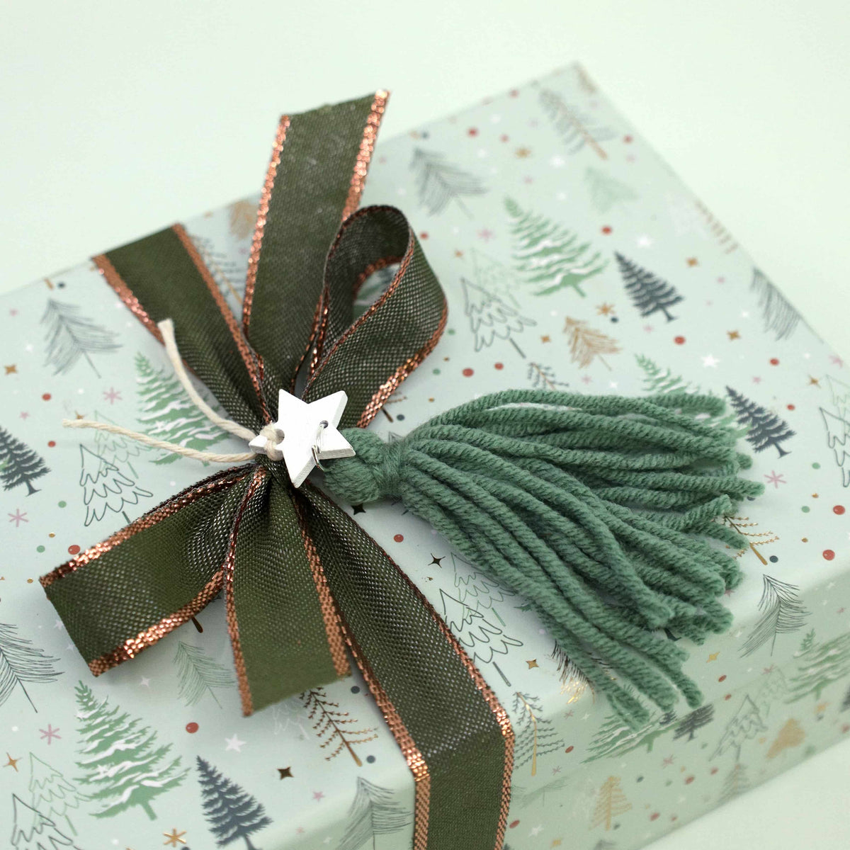 Un cadeau emballé dans du papier bleu clair orné de motifs d'arbres, noué avec un ruban vert et cuivré, agrémenté d'un pompon en fil vert et d'une petite breloque en forme d'étoile argentée, est parfaitement mis en valeur par l'étiquette « Geschenkanhänger Tannenbaum » d'Artoz Papier AG — idéale pour apporter cette touche spéciale « DIY-Projekte ».