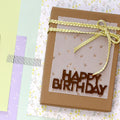 Une boîte à surprise Artoz Papier AG Pure A6 grocer kraft avec un panneau HAPPY BIRTHDAY doré et un ruban tressé jaune repose sur des papiers floraux pastel, créant ainsi un charmant Geschenkverpackung.