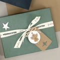 La Geschenk-Box Pure A6 d'Artoz Papier AG en moos grün est enveloppée d'un ruban portant la mention "frohe WEIHNACHTEN" et décorée d'une étoile, d'une étiquette marron et d'un ange en bois.
