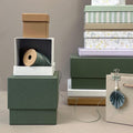 Une pile d'Artoz Papier AG Geschenk-Box Pure Grösse L en vert mousse, avec une bobine de ficelle verte et un petit éventail en papier accroché à un sac cadeau beige, accentué par des couleurs sourdes et des motifs.