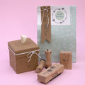 La Geschenk-Box Pure Grösse M Kraft d'Artoz Papier AG est présentée avec des tampons en forme de cœur, des nœuds en ficelle et des motifs floraux. Des tampons botaniques en bois sont disposés devant, le tout sur un fond rose pastel.