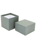 Une boîte à geschenk simple, carrée, vert mousse Pure Grösse M d'Artoz Papier AG avec son couvercle mis de côté, montrant l'intérieur blanc vide.