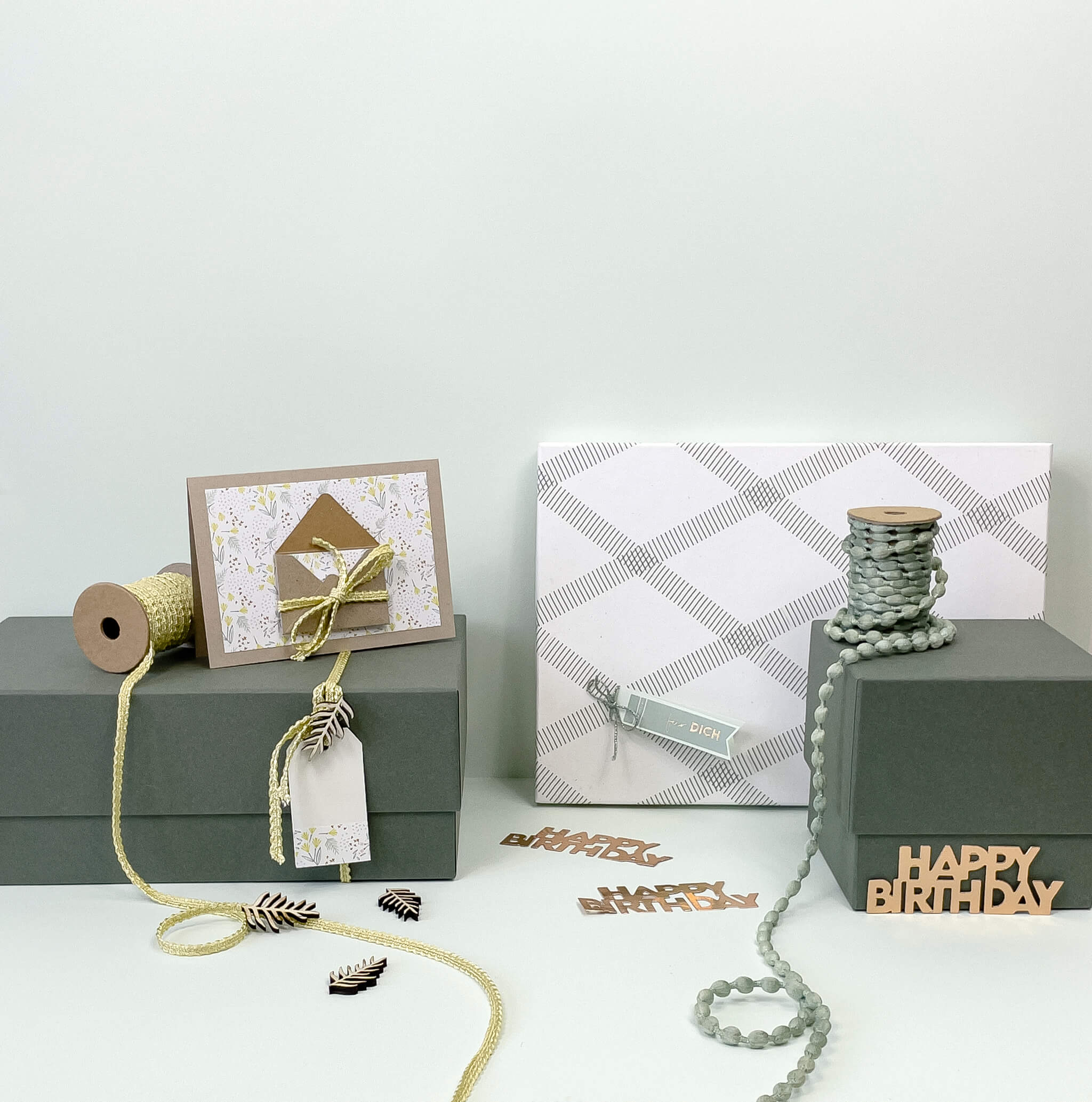Deux boîtes-cadeaux vertes « Pure L » rectangulaires de la marque Artoz Papier AG (couleurs variées) sont disposées avec soin sur une surface claire, devant un fond blanc, accompagnées de bobines de ficelle, d’étiquettes décoratives, d’une carte d’anniversaire, d’un cadeau emballé et de découpes en bois portant l’inscription « Happy Birthday » — mettant ainsi en valeur un emballage cadeau élégant, idéal pour toutes les occasions.