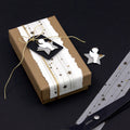 L'Artoz Papier AG Geschenk-Box Pure S rechteckig grocer kraft, enveloppée dans un papier blanc à motif d'étoiles avec une ficelle dorée et une étiquette noire à étoiles représentant un ange blanc, est idéale pour les stilvolles Geschenke verpacken. Des ciseaux et un ruban à étoiles dorées complètent l'ensemble.