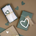 Trois Geschenk-Box Pure S rechteckig moos grün d'Artoz Papier AG et deux boîtes brunes Buchkarton sont ornées de formes de cœur, de nœuds en ficelle, de papier de soie blanc et d'étiquettes en forme de cœur éparpillées, comme autant de délicieux accents d'emballage cadeau.