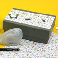 Une Geschenk-Box Pure M d'Artoz Papier AG aux motifs floraux et papillons est posée sur une surface jaune à côté d'un dévidoir de ruban correcteur, d'un stylo noir et d'un papier à fleurs assorti.