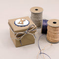 Une Artoz Papier AG Geschenk-Box Pure Grösse XS kraft est posée à côté de trois bobines de ficelle décoratives. La petite boîte brune avec un cercle festonné et un couvercle découpé en forme de lapin est terminée par de la ficelle, présentée sur une surface claire.