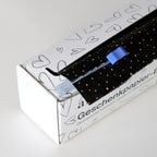 Une boîte de papier cadeau Artoz Papier AG, comprenant du papier cadeau de différentes couleurs — une boîte distributrice rechargeable — contient un rouleau de papier cadeau noir à pois dorés ainsi qu'un cutter en plastique bleu, qui se range facilement sur le papier. La boîte blanche est décorée de motifs en forme de cœur et comporte un texte en allemand.