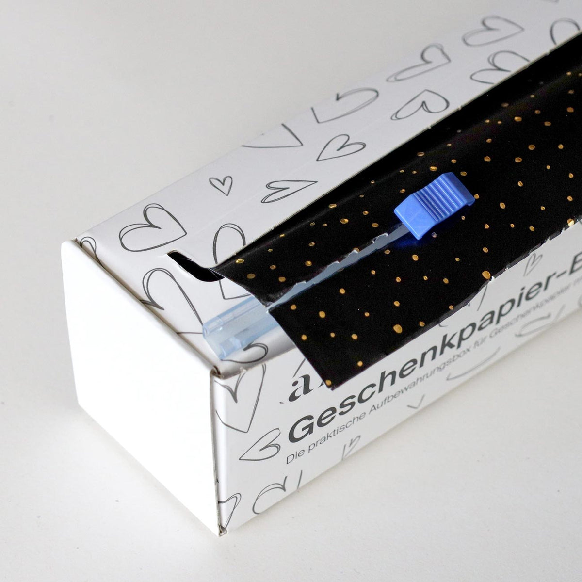 Une boîte de papier cadeau Artoz Papier AG, comprenant du papier cadeau de différentes couleurs — une boîte distributrice rechargeable — contient un rouleau de papier cadeau noir à pois dorés ainsi qu'un cutter en plastique bleu, qui se range facilement sur le papier. La boîte blanche est décorée de motifs en forme de cœur et comporte un texte en allemand.