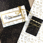 Trois cadeaux emballés arborent des motifs de cœurs et de pois blancs, noirs et dorés issus du rouleau de papier cadeau « Geschenkpapier Rolle », conçu pour s'adapter à la boîte de recharge « Geschenkpapier Nachfüllbox » d'Artoz Papier AG. Chacun est noué avec une ficelle dorée, et l'une des étiquettes porte l'inscription « öffnen UND FREUEN ! » en allemand. Les cadeaux sont disposés sur du papier cadeau Artoz assorti.
