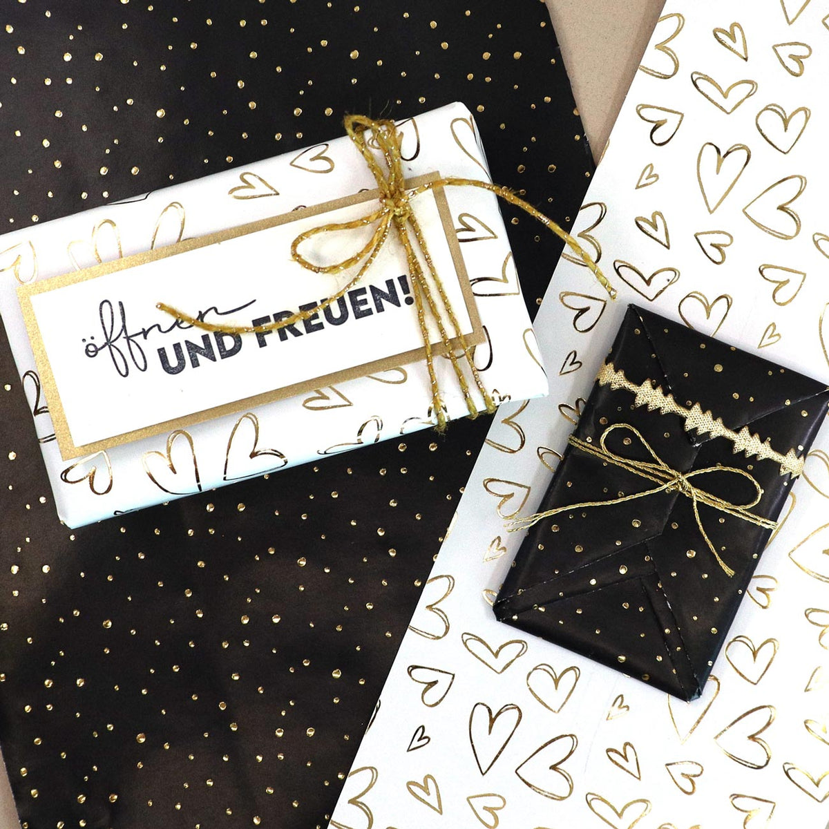 Trois paquets cadeaux mettant en valeur la boîte « Geschenkpapier Box » d’Artoz Papier AG, comprenant du papier cadeau de différentes couleurs — boîte distributrice rechargeable, décorée de papier cadeau doré et blanc orné de cœurs et de pois. Deux d'entre eux sont noués avec des rubans dorés. Une étiquette porte l'inscription « öffnen UND FREUEN ! », ce qui signifie « ouvrez ET RÉJOUISSEZ-VOUS ! » en allemand.