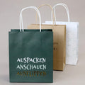 Trois sacs Artoz Papier AG Geschenktaschen-Set "Auspacken - Anschauen - Ausflippen" sont disposés : un sac vert avec un texte en allemand sur le devant, un sac Kraftpapier marron simple au milieu et un sac blanc avec des points et des feuilles dorés à l'arrière.
