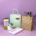 Geschenktaschen-Set "Du bist wundervoll" von Artoz Papier AG liegt mit grünen Polka-Dot-Taschen, Papiertüten mit Griff, Karte, verpackten Geschenken, Konfetti und Blumen dekorativ auf lilafarbenem Hintergrund.