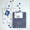 Deux sacs cadeaux Artoz Papier AG Geschenktaschen-Set "Happy Birthday" (330) avec des poignées blanches sont présentés - l'un avec un motif floral bleu et une souris, l'autre bleu foncé avec des abeilles et "Jedes Jahr ein neues Abenteuer". Des confettis bleus et blancs les entourent.