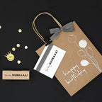 Le set Geschenktaschen Happy Birthday (317) d'Artoz Papier AG comprend un sac cadeau en papier kraft avec des motifs d'anniversaire et de ballon, une carte HURRAAA ! assortie, un tampon, des confettis dorés et un autocollant smiley couronné sur fond noir.