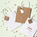 Le set Artoz Papier AG Geschenktaschen-Set Happy Birthday (369) comprend trois Kraftpapier-Partytüten en forme de cœur avec Griff et le motif "Happy Birthday", chacun avec une étiquette attachée, disposés au milieu de confettis dorés et blancs sur une surface claire.