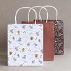 Drei Geschenktaschen aus dem „Geschenktaschen-Set Weihnachten 3er-Set“ von Artoz Papier AG mit gedrehten weißen Tragegriffen sind in einer Reihe angeordnet. Die vordere Tasche ist mit einem weißen Weihnachtsmotiv verziert, die mittlere ist einfarbig rostrot und die hintere zeigt ein Blumenmuster – alles vor einem neutralen beigen Hintergrund.