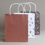 Drei Geschenkbeutel aus dem Geschenktaschen-Set „Weihnachten 3er-Set“ von Artoz Papier AG mit weißen Papier-Tragetaschen und Drehgriff stehen in einer Reihe: Die vordere Geschenktasche aus Kraftpapier ist braun mit kleinen goldenen Punkten, die mittlere ist weiß mit verschiedenen kleinen Illustrationen und die hintere ist schwarz mit einem hübschen Blumenmuster gestaltet.