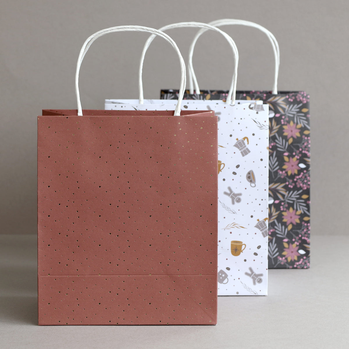 Drei Geschenkbeutel aus dem Geschenktaschen-Set „Weihnachten 3er-Set“ von Artoz Papier AG mit weißen Papier-Tragetaschen und Drehgriff stehen in einer Reihe: Die vordere Geschenktasche aus Kraftpapier ist braun mit kleinen goldenen Punkten, die mittlere ist weiß mit verschiedenen kleinen Illustrationen und die hintere ist schwarz mit einem hübschen Blumenmuster gestaltet.