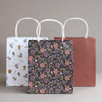 Drei Geschenktüten aus Papier stehen nebeneinander: eine weiße mit Illustrationen zum Thema Weltraum, eine schwarze mit rosa Blumenmustern und eine kastanienbraune mit kleinen goldenen Punkten – alle Teil des „Geschenktaschen-Set Weihnachten 3er-Set“ von Artoz Papier AG mit weißen Tragetaschen mit gedrehten Henkeln –, angeordnet vor einem neutralen Hintergrund.