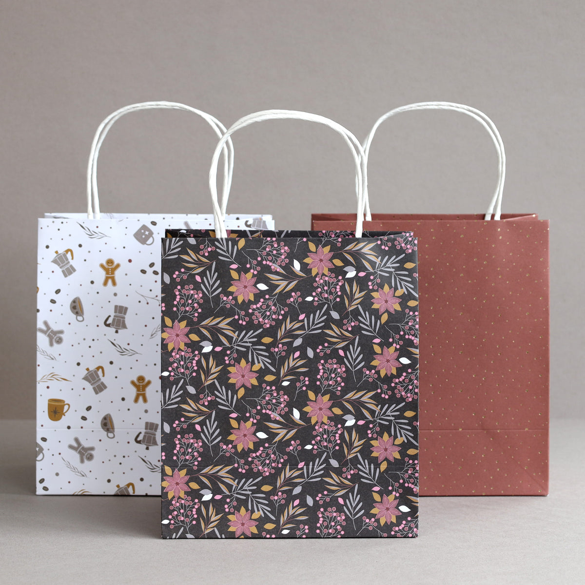 Drei Geschenktüten aus Papier stehen nebeneinander: eine weiße mit Illustrationen zum Thema Weltraum, eine schwarze mit rosa Blumenmustern und eine kastanienbraune mit kleinen goldenen Punkten – alle Teil des „Geschenktaschen-Set Weihnachten 3er-Set“ von Artoz Papier AG mit weißen Tragetaschen mit gedrehten Henkeln –, angeordnet vor einem neutralen Hintergrund.
