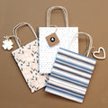 Drei Geschenktaschen-Set mit Pflanzenmustern (333) der Artoz Papier AG mit Henkeln in Blumen-, Punkt- und Streifendesign liegen auf einer braunen Oberfläche; zwei haben dekorative Anhänger in Form eines Klees und eines Herzens.