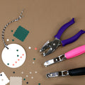 Ein runder weißer Anhänger mit herzförmigen Löchern, umgeben von farbigen Papierformen und drei Artoz Papier AG Handlocher Herz mit lila, rosa und schwarzen Griffen auf einer braunen Oberfläche.