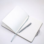 Un carnet A5 ouvert et personnalisable, à quadrillage, de la marque Artoz Papier AG, de couleur beige, avec votre nom ou le texte de votre choix, repose sur un fond blanc ; à côté se trouvent un stylo à bille argenté assorti et un ruban marque-page en tissu beige.