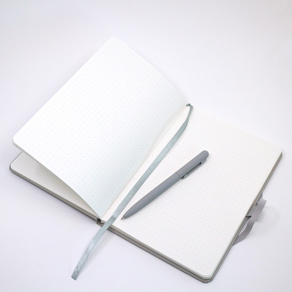 Un carnet A5 ouvert et personnalisable, à quadrillage, de la marque Artoz Papier AG, de couleur beige, avec votre nom ou le texte de votre choix, repose sur un fond blanc ; à côté se trouvent un stylo à bille argenté assorti et un ruban marque-page en tissu beige.