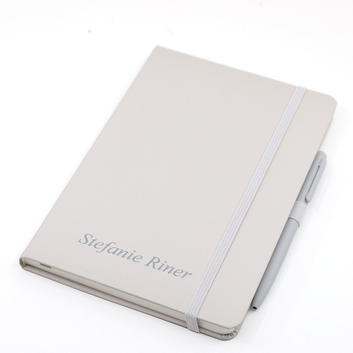 Un carnet A5 beige à couvercle fermé avec stylo, de la marque Artoz Papier AG, personnalisé au nom de Stefanie Riner sur la couverture, fermé par un élastique et muni d'un stylo à bille beige assorti fixé sur le côté.
