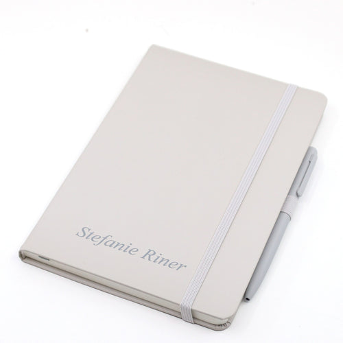 Carnet A5 beige avec le nom/texte Stefanie Riner sur la couverture, maintenu par un élastique et accompagné d'un stylo à bille assorti.