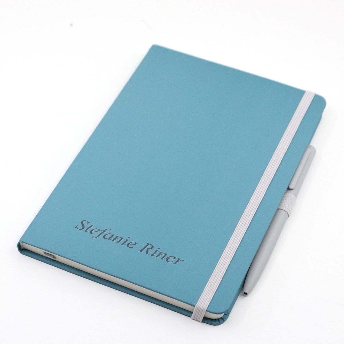 Ein türkisfarbenes A5-Notizbuch mit Kugelschreiber von Artoz Papier AG, personalisiert mit „Stefanie Riner“ auf dem Einband. Ein weißes Gummiband hält das personalisierte Notizbuch zusammen und befestigt den weißen Kugelschreiber an der rechten Seite. Das Produkt liegt auf weißem Hintergrund.