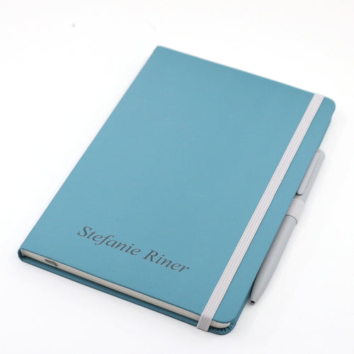 Carnet A5 bleu avec nom/texte, muni d'un élastique, d'un stylo et du nom Stefanie Riner gravé sur la couverture.