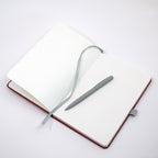 Un carnet A5 personnalisable avec stylo de la marque Artoz Papier AG, dans une élégante teinte bordeaux et orné du nom ou du texte de votre choix, est présenté avec des pages pointillées, un ruban marque-page gris et un stylo gris assorti disposé sur ses pages immaculées, le tout sur un fond blanc épuré.