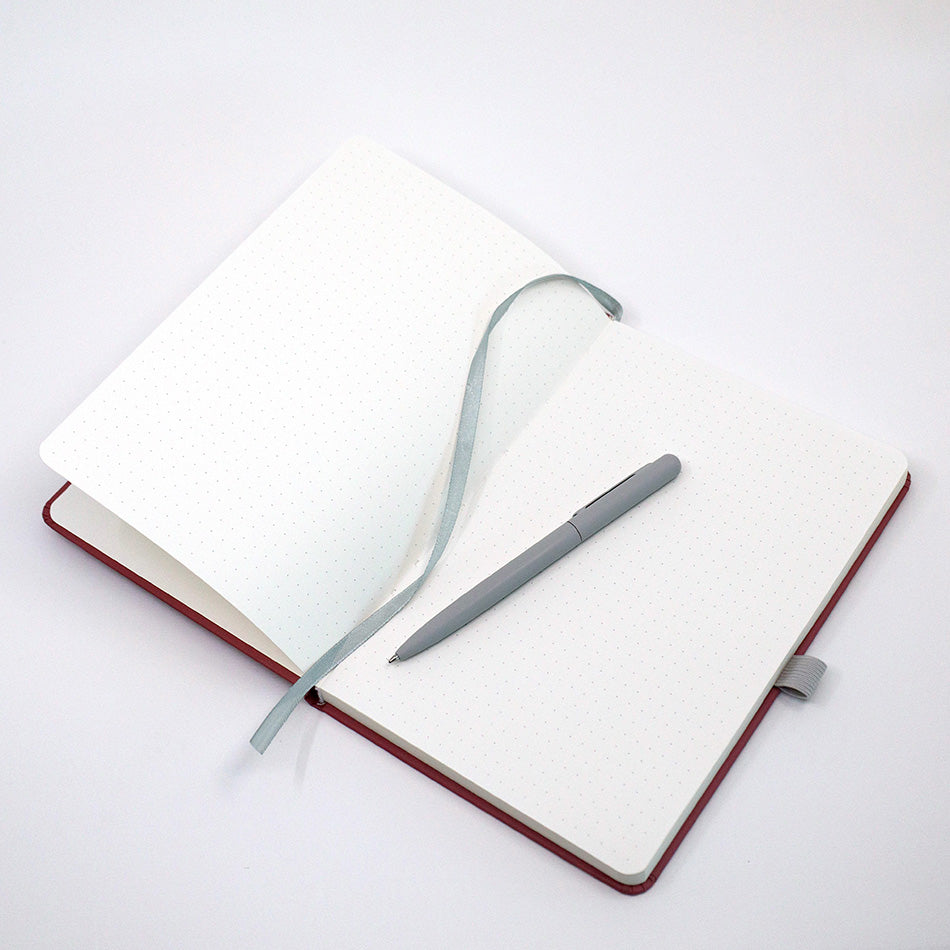 Un carnet A5 personnalisable avec stylo de la marque Artoz Papier AG, dans une élégante teinte bordeaux et orné du nom ou du texte de votre choix, est présenté avec des pages pointillées, un ruban marque-page gris et un stylo gris assorti disposé sur ses pages immaculées, le tout sur un fond blanc épuré.