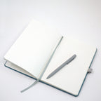 Ein offenes A5-Notizbuch mit Kugelschreiber, türkis, mit deinem Namen/Text von Artoz Papier AG, mit einem grauen Lesezeichenband, auf dessen gepunkteten Seiten ein personalisierter Kugelschreiber ruht – das Ganze vor einem schlichten weißen Hintergrund.