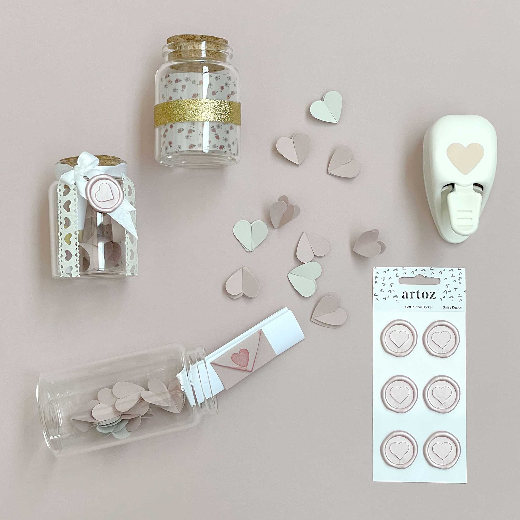 Une présentation à plat des fournitures de bricolage d'Artoz Papier AG sur une surface beige clair, idéale pour Basteln ou Karten gestalten, comprenant des pots avec des cœurs, des cœurs en papier éparpillés, la perforatrice Stanzer S Herz et des autocollants ronds en forme de cœur.